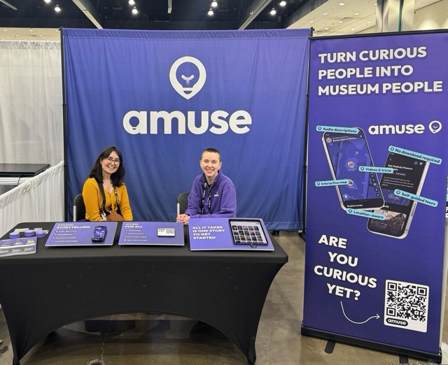 Two Amuse team members at their exhibition booth at the American Alliance of Museums conference, with a banner reading 'Turn curious people into museum people.'