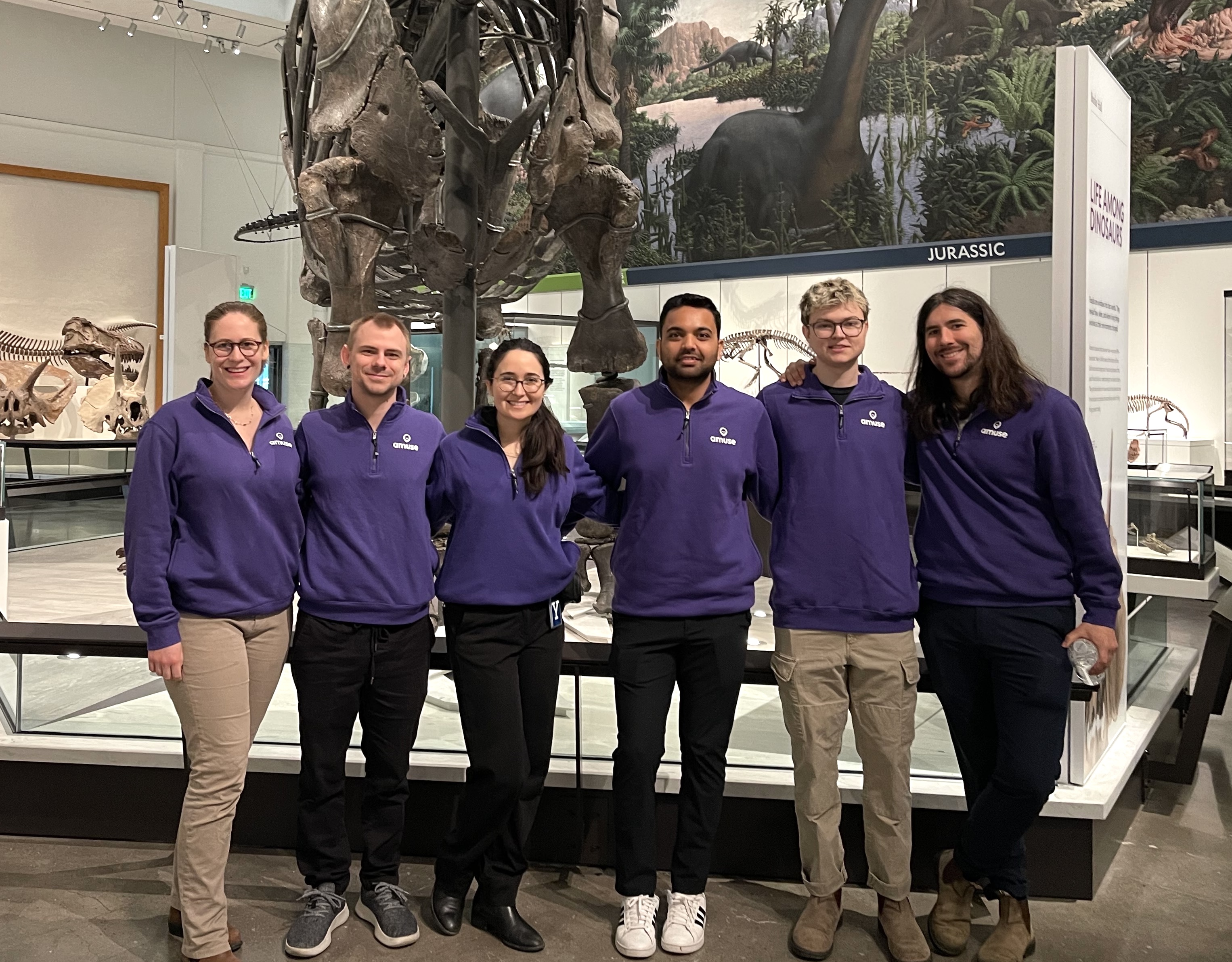 The Amuse team in matching purple sweatshirts standing in the Jurassic hall of the Yale Peabody Museum, surrounded by dinosaur skeletons.